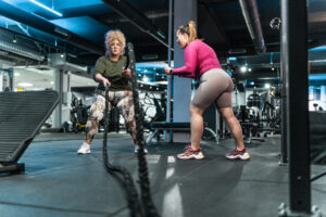 Two women engaged in a personal training session using battle ropes in a gym.