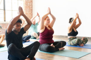 Yoga class participants in seated meditation pose with hands above their heads.